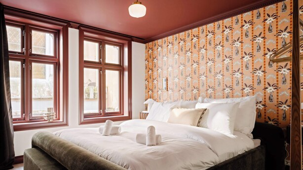 A neatly made bed with white linens and two towels, a wooden headboard, and a window with curtains.