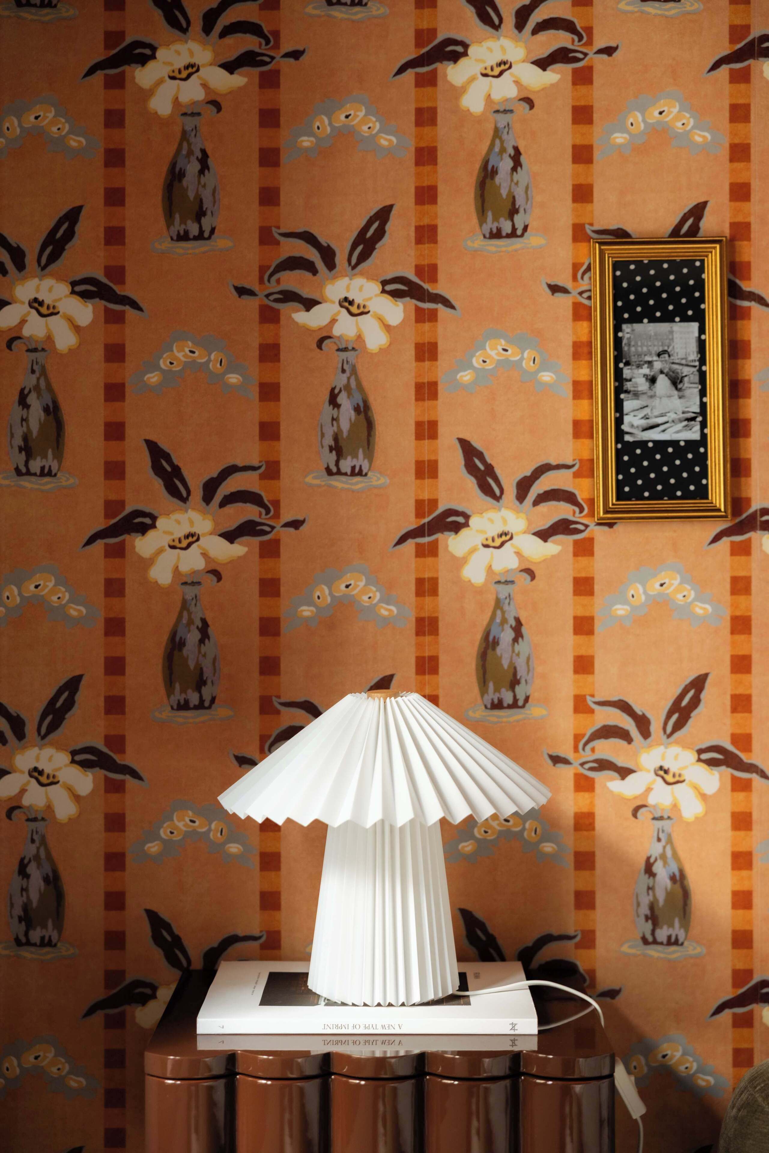 Una stanza con carta da parati decorata, una lampada bianca su un tavolo e un quadro appeso al muro.