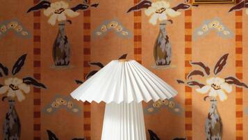 Una stanza con carta da parati decorata, una lampada bianca su un tavolo e un quadro appeso al muro.
