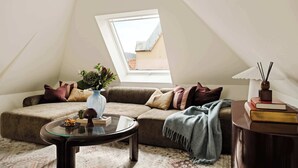 Ein gemütliches Schlafzimmer im Loft-Stil mit schrägem Dach, einem großen Fenster, einer Couch, einem Glastisch und einem Nachttisch mit Büchern und einer Lampe.