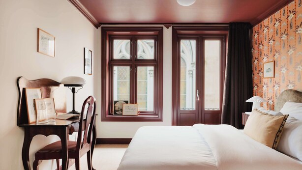 Ein Schlafzimmer mit einem großen Bett, einem Schreibtisch mit Stuhl, zwei Fenstern mit Vorhängen und einer Wand mit Muster.