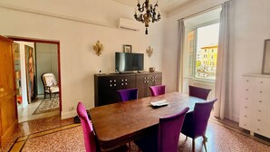 Uma área de refeições com uma mesa de madeira e cadeiras roxas, um armário de madeira escura com uma televisão, e vista para a cidade através de uma janela grande.