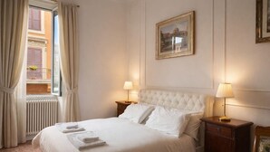 Cama bem arrumada com lençóis brancos e duas almofadas, janela com cortinas, radiador e um quadro na parede.