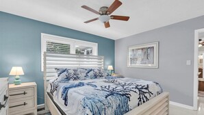 Une chambre à coucher avec un lit, un ventilateur de plafond, une fenêtre avec des stores, une table de chevet avec une lampe et un tableau encadré au mur.
