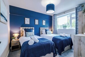 Una habitació amb dos llits, una nitòria amb una làmpada, una finestra amb cortines i quadres penjats a la paret.