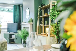 Een woonkamer met een televisie, groene gordijnen, een boekenkast met planten, en een eethoek met een tafel en stoelen.