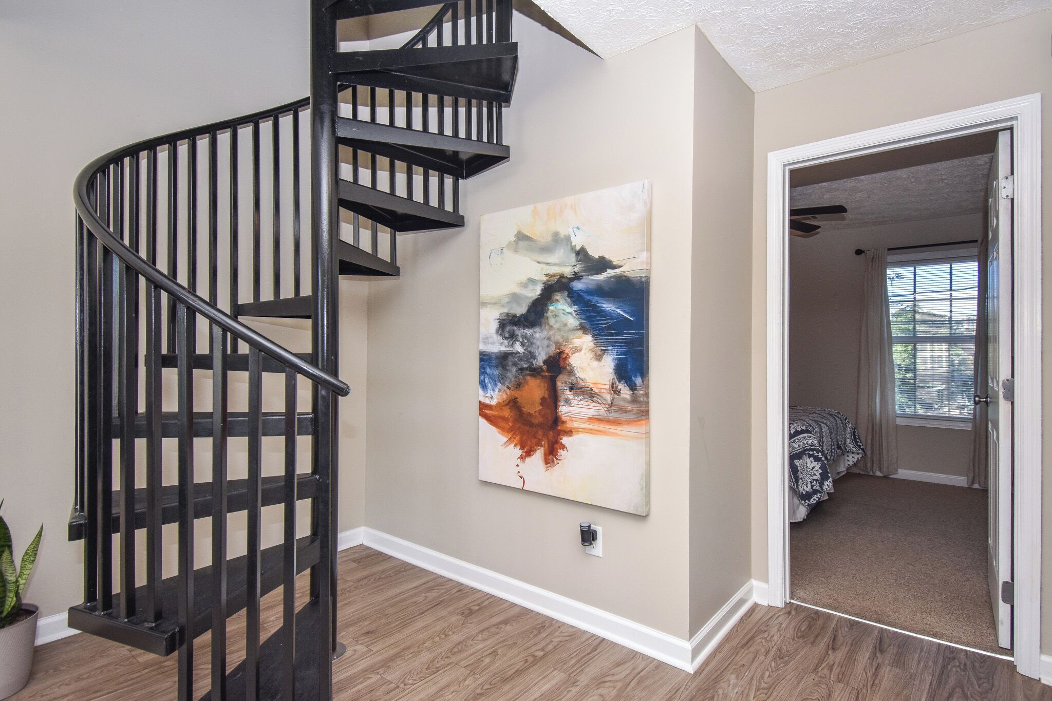 A spiral staircase with a modern design, a large abstract painting on the wall, and a doorway leading to a bedroom with a window.