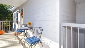 A covered outdoor area with a white railing, a small round table, two chairs with blue cushions, and a vase with flowers.