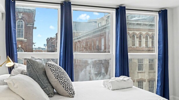 A hotel room with a large bed, blue curtains, and a view of historic buildings. - Posh Center Spot: 2BR Fits 6 Guests Near Sights (Philadelphia)