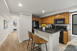 A modern kitchen with wooden cabinets, a black refrigerator, a microwave, and a central island with bar stools. - Prime 2BR Sleeps 6 Oasis | Near The Liberty Bell (Philadelphia)
