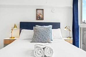Una cama bien hecha con toallas enrolladas, un cabecero y dos lámparas de noche.