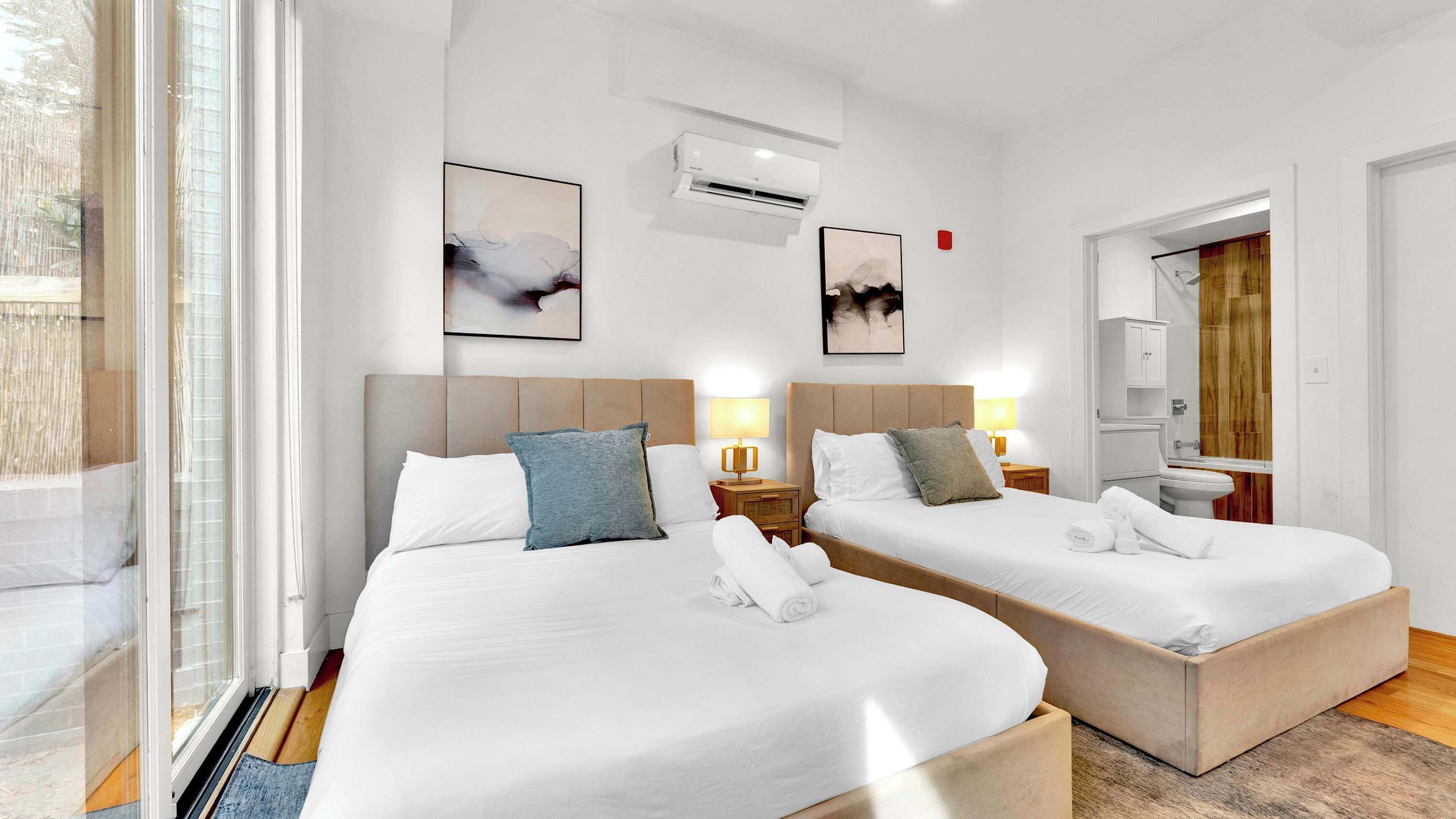 Ein Hotelzimmer mit zwei Betten, einem Nachttisch mit Lampe, einer Wandklimaanlage und einem großen Fenster.