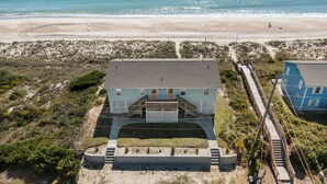 A beachfront property with a prominent house featuring a large porch and a staircase leading to the entrance. - Classic Oceanfront Beach Cottage! (Emerald Isle)