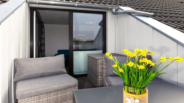 A balcony with grey sofas, a table, and a vase of yellow flowers. - Ferienwohnung Balkon zur Sonnenseite in Bensersiel (Bensersiel)