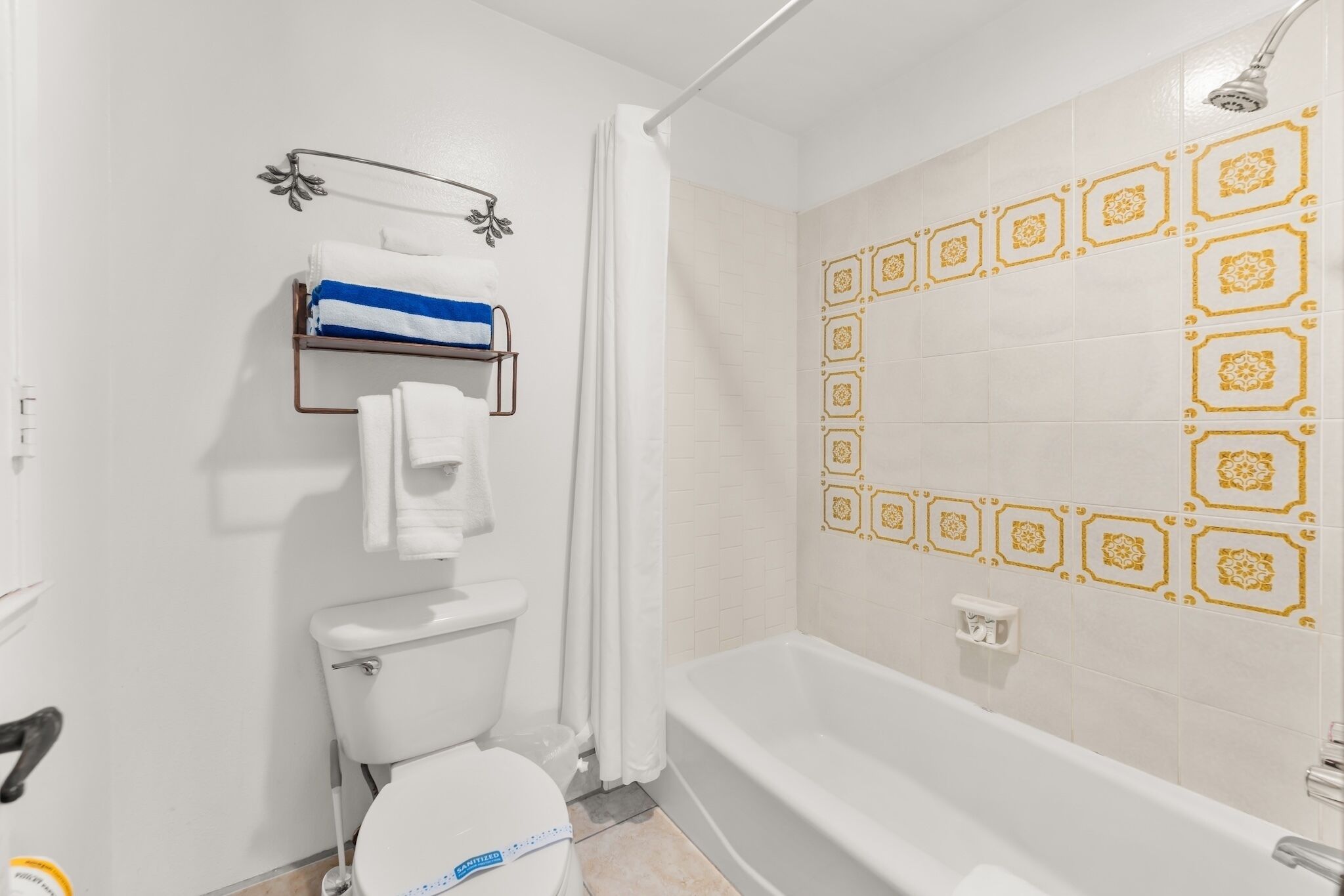 Een badkamer met toilet, douche en bad. Er is een handdoekhouder met een handdoek en een blauw-wit gestreepte handdoek. De wanden zijn betegeld met decoratieve tegels met een geometrisch patroon.