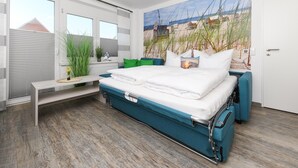 A modern bedroom with a large bed, a window with blinds, a bench, and a beach-themed wall mural.