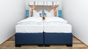 Cama de casal com roupa de cama branca e base estofada em azul, um dossel listrado e duas luminárias fixadas na parede.