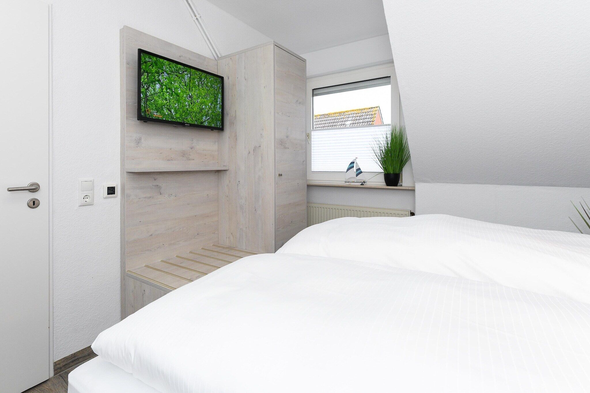 A modern bedroom with a bed, a mounted TV, a window with a view of a building, and a potted plant on a shelf.