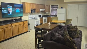 Une cuisine compacte avec des meubles en bois, un four à micro-ondes, une cuisinière et un réfrigérateur. Une table à manger avec des chaises est placée au centre, et un canapé violet est adossé au mur.
