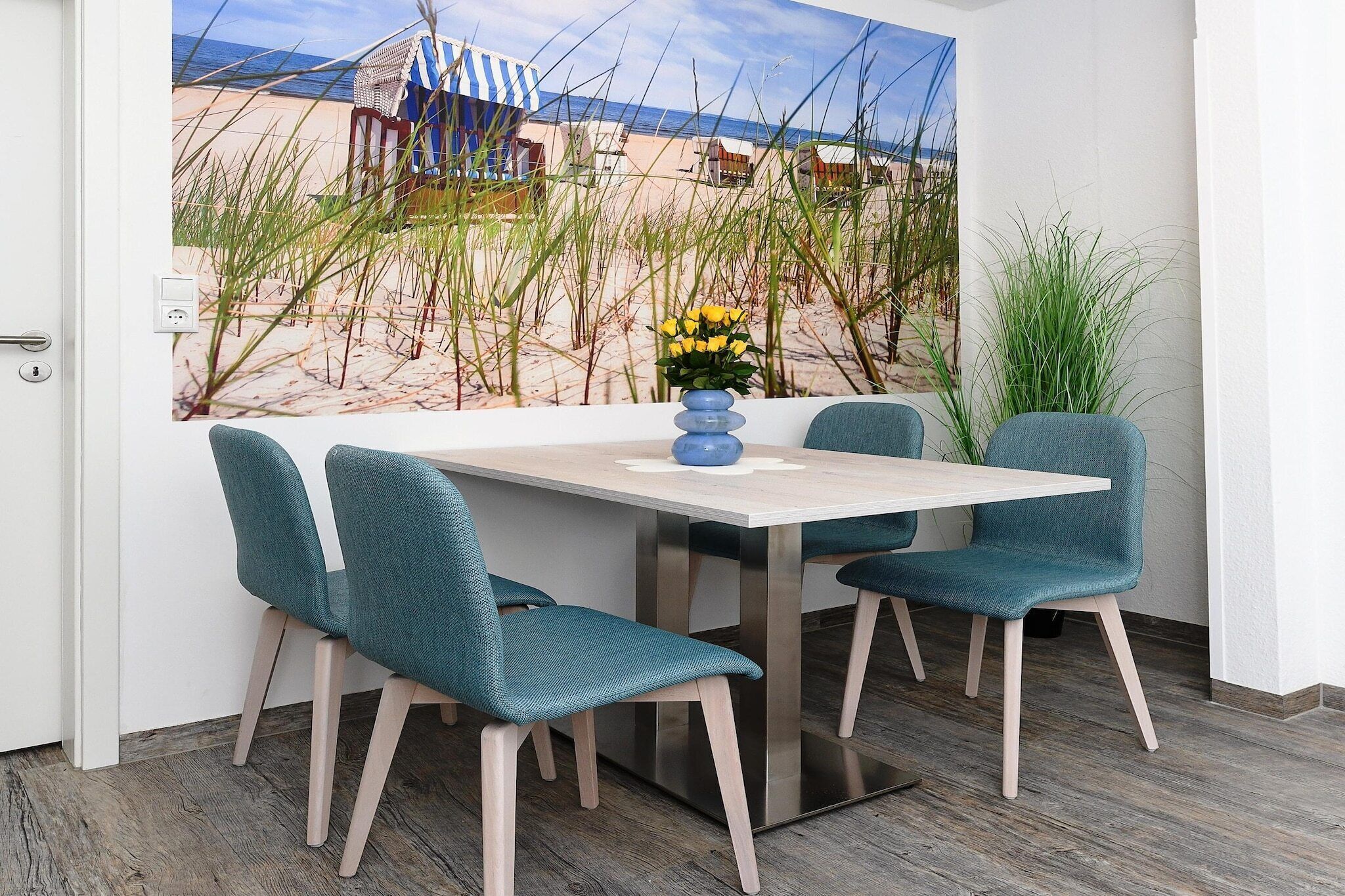 A dining area with a beach view, a table, four teal chairs, and a vase with yellow flowers.