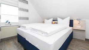 Un lit bien fait, avec du linge de lit blanc, une fenêtre avec des rideaux, une petite décoration représentant un voilier et une table de chevet avec une lampe.