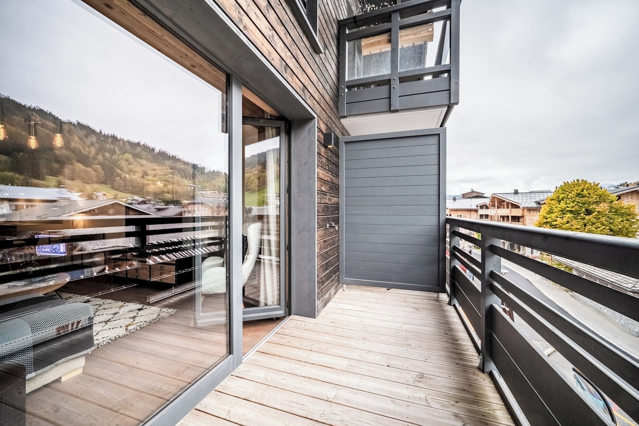 Ein moderner Balkon mit Holzboden und Glastüren, mit Blick auf ein Wohngebiet mit Häusern und einen bewaldeten Hügel.