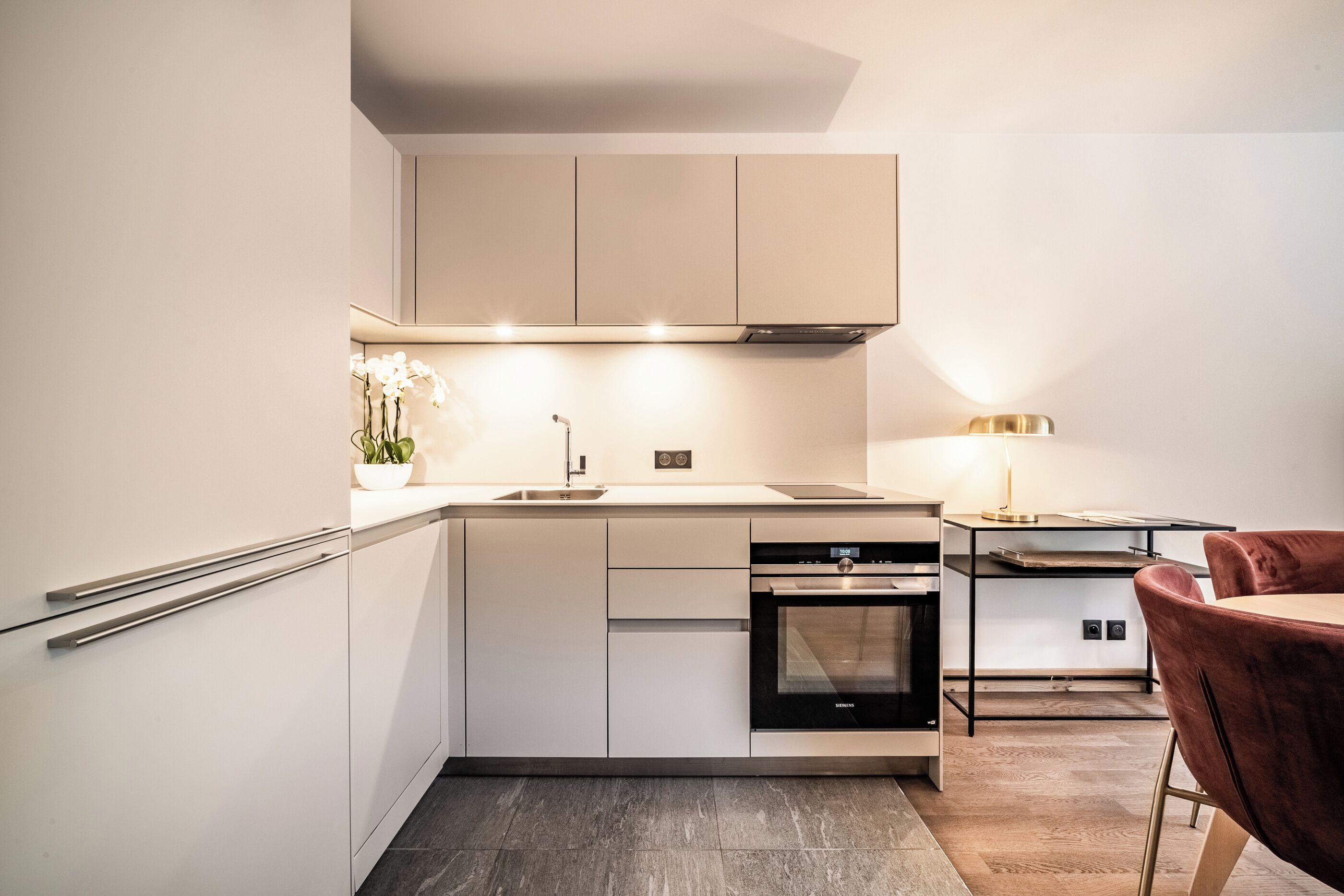 Een moderne keuken met witte kasten, een ingebouwde oven en een eethoek met een rode stoel en een lamp.