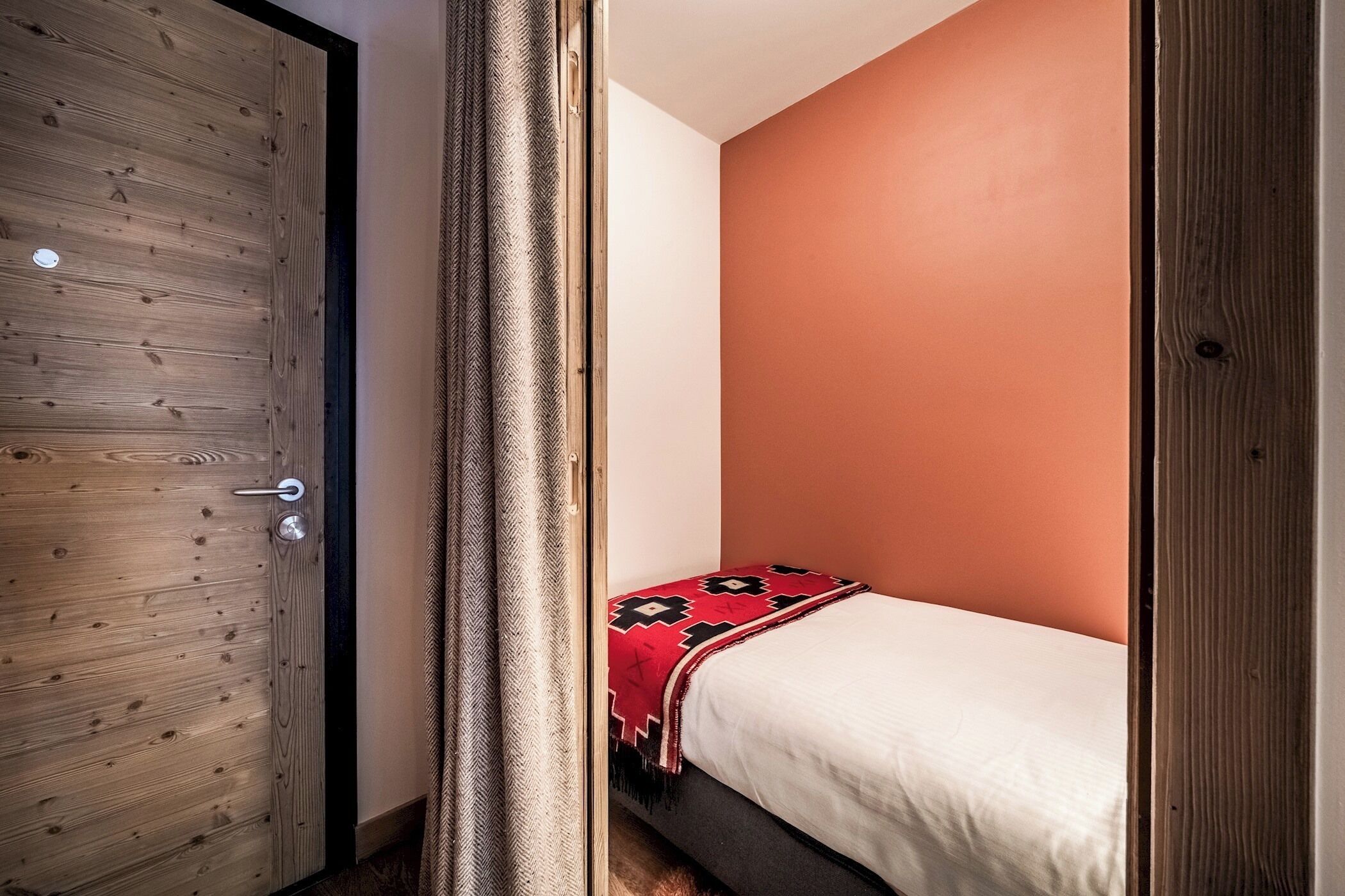 Une pièce avec une porte en bois, un lit recouvert d’une couverture à motifs rouges et noirs, et un mur de couleur orange unie.
