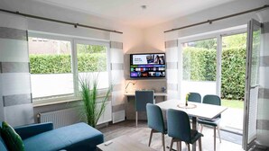A modern living room with a TV mounted on the wall, a dining area with chairs and a table, and a sofa with a remote control on it. - Moderne Ferienwohnung mit Terrasse in ruhiger Lage (Bensersiel)