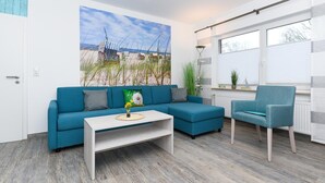 A modern living room with a blue sofa, a white coffee table, and a blue armchair. There is a large beach-themed painting on the wall and a window with blinds. - Moderne Ferienwohnung mit Terrasse in ruhiger Lage (Bensersiel)