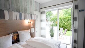 A bedroom with a large bed, a wooden headboard, a nightstand, and a view of the garden through a sliding glass door. - Moderne Ferienwohnung mit Terrasse in ruhiger Lage (Bensersiel)