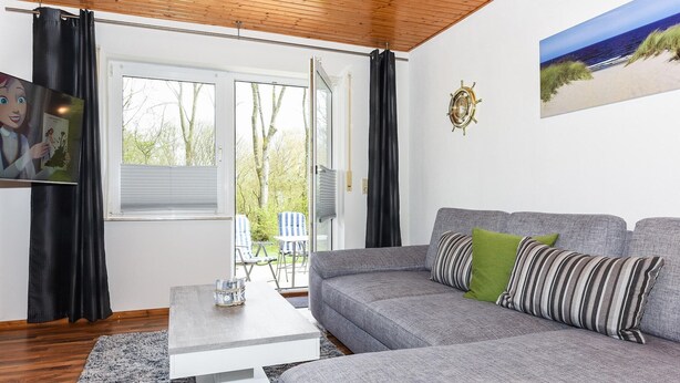A modern living room with a grey sofa, a coffee table, and a view of the outdoors through a large window. - Geräumige Ferienwohnung mit Terrasse in Bensersiel (Bensersiel)