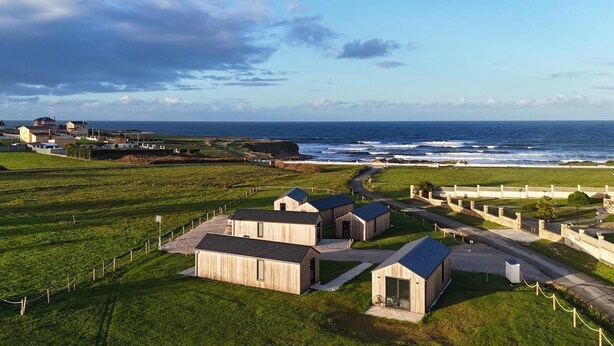 A coastal area with wooden buildings, a fenced pathway, and a view of the ocean. - Cabañas Camiño do Mar  (Foz)