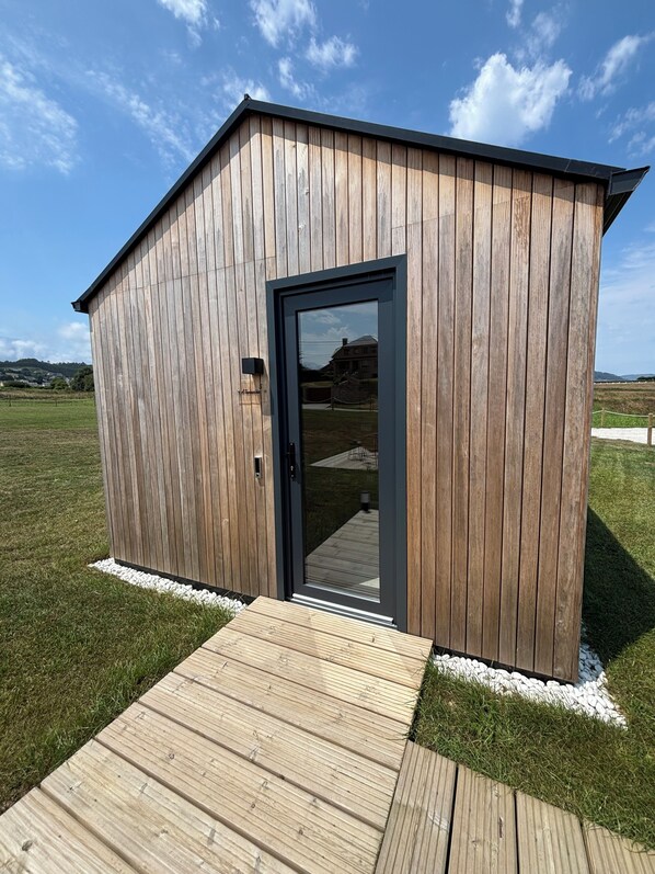 A wooden shed with a glass door, a wooden deck, and a grassy field in the background. - Cabañas Camiño do Mar (Foz)
