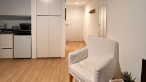 Un espacio habitable compacto con kitchenette, un sillón blanco y un pequeño mueble para televisión.