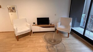 Una sala de estar moderna con piso de madera, una mesa de centro de vidrio y dos sillones blancos. Un televisor de pantalla plana está sobre una consola blanca.