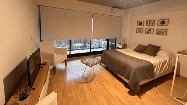 Un dormitorio moderno con cama, escritorio, silla y una pequeña mesa. Se ve una ventana con vistas a muebles de exterior.
