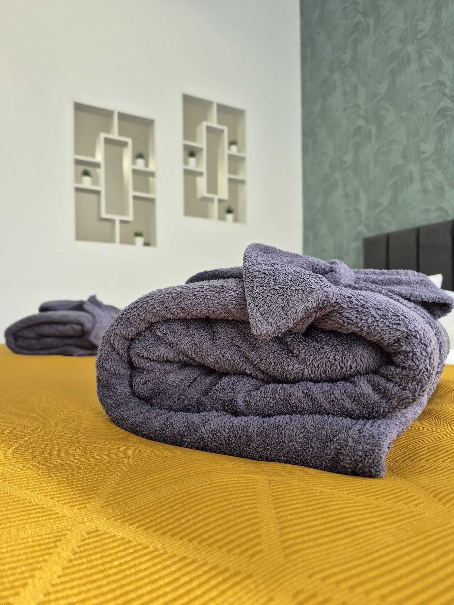 Une serviette grise, soigneusement pliée, est posée sur un couvre-lit jaune à motifs dans une pièce dont le mur présente un décor géométrique et où la tête de lit est de couleur sombre.