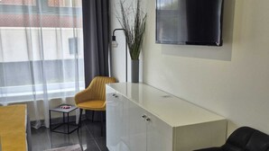 Un salon moderne avec un canapé, un fauteuil noir, un téléviseur fixé au mur, une petite table d’appoint et une plante en pot.