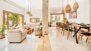 Amplia sala de estar con sofá, mesa de comedor y sillas. Se observa un pilar de piedra, un banco de madera y una planta en maceta.