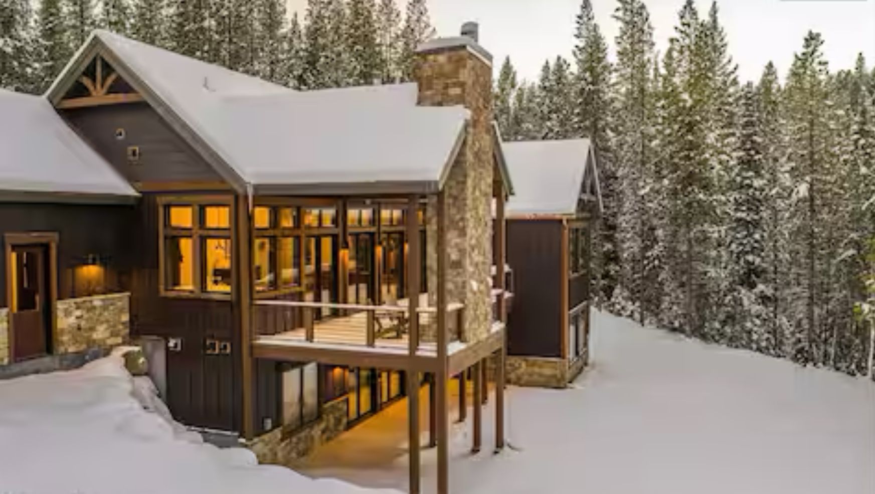Une grande cabane en bois à plusieurs niveaux, dotée d’une cheminée en pierre, entourée d’arbres couverts de neige.