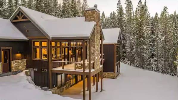 Una cabaña grande de madera, de varios niveles, con una chimenea de piedra, rodeada de árboles cubiertos de nieve.