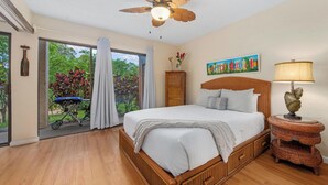 Een slaapkamer met een houten bed, een plafondventilator, een raam met gordijnen en uitzicht op groen.