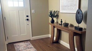A hallway with a wooden floor, a rug, a door, a small table with a vase and figurines, and a framed picture on the wall.
