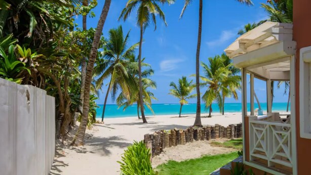 Vista de la playa con palmeras, una valla blanca y un edificio con porche.