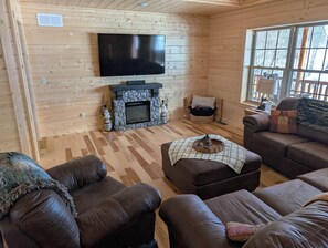 A cozy cabin interior with a stone fireplace, a flat-screen TV mounted on the wall, and leather sofas arranged around a central ottoman.