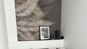 Ein modernes Wohnzimmer mit einem beigefarbenen Sofa, zwei dunkelblauen Kissen, einer grauen Decke, einem schwarzen Couchtisch mit einer dekorativen Vase und Fernbedienung sowie einem gerahmten Bild an der Wand.