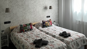 Ein Schlafzimmer mit zwei Betten, blumigem Bettbezug, Deckenventilator, Fenster mit Vorhängen und einem kleinen Nachttisch mit Lampe.