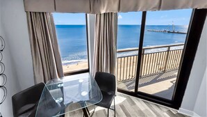 Une pièce avec une table en verre et deux chaises, de grandes fenêtres donnant sur la plage et une jetée, ainsi qu’un balcon doté d’une balustrade.