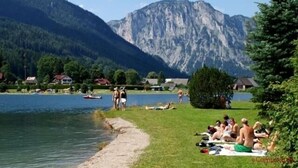 A lakeside view with people sunbathing on the grass, a boat on the water, and mountains in the background. - Becci`s Apartment- Free Parking (Kapfenberg)